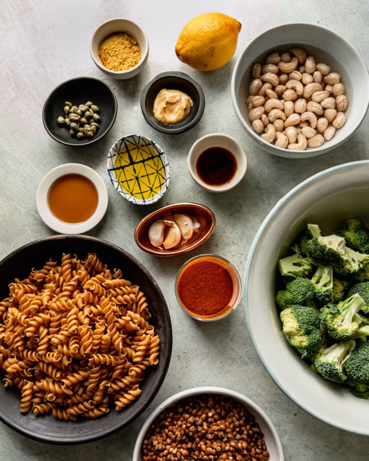 An overhead shot shows ingredients needed for a vegan broccoli Caesar pasta salad recipe. Most ingredients are measured out in small bowls.
