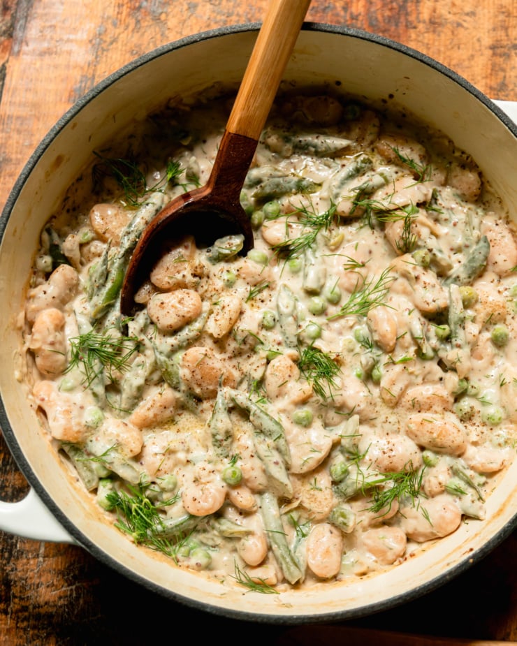 An overhead shot shows a braiser pot filled with creamy dilly beans and peas. A serving spoon sticks out of the pot.