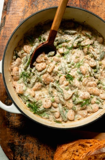 An overhead shot shows a braiser pot filled with creamy dilly beans and peas. A serving spoon sticks out of the pot and some bread is seen to the side.