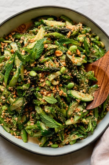An up close, overhead shot shows a round ceramic dish filled with a roasted asparagus salad. The salad also features shelled edamame, snap peas, cucumber, fresh mint, and ginger lime dressing. The salad is dusted with finely chopped salted peanuts. A wooden serving spoon is tucked into the salad.