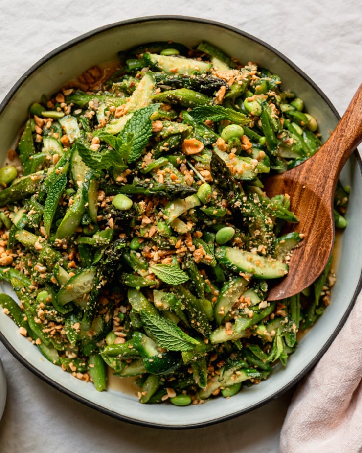 An up close, overhead shot shows a round ceramic dish filled with a roasted asparagus salad. The salad also features shelled edamame, snap peas, cucumber, fresh mint, and ginger lime dressing. The salad is dusted with finely chopped salted peanuts. A wooden serving spoon is tucked into the salad.