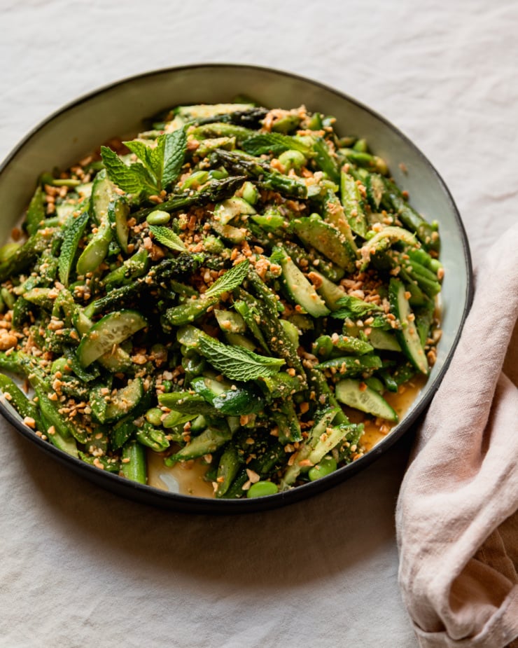 A 3/4 angle shot shows a round ceramic dish filled with a roasted asparagus salad. The salad also features shelled edamame, snap peas, cucumber, fresh mint, and ginger lime dressing. The salad is dusted with finely chopped salted peanuts.