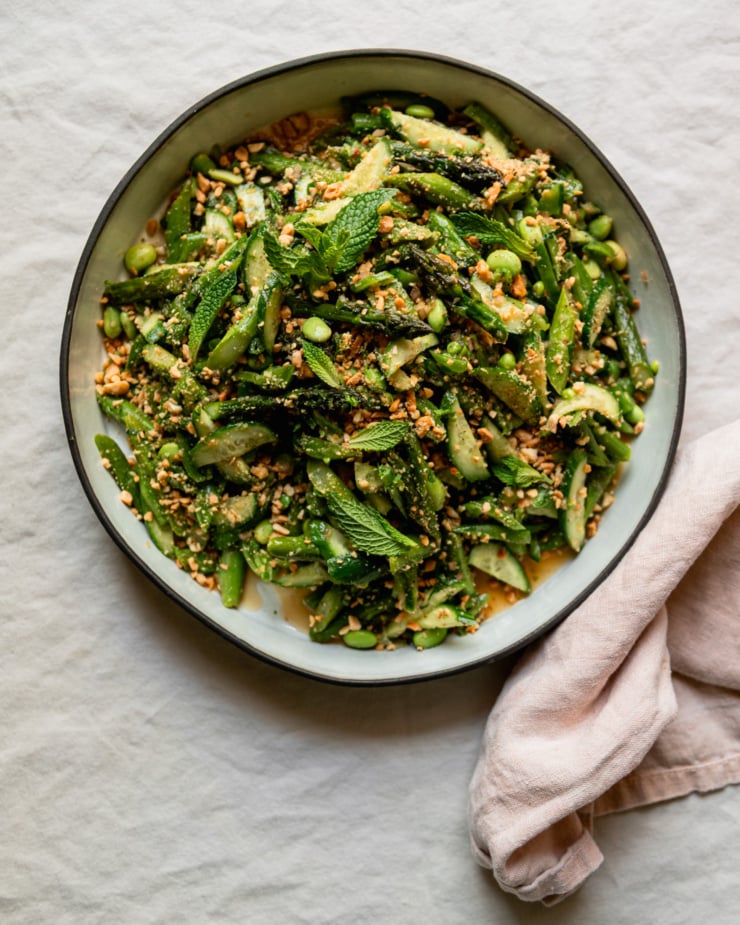 An overhead shot shows a round ceramic dish filled with a roasted asparagus salad. The salad also features shelled edamame, snap peas, cucumber, fresh mint, and ginger lime dressing. The salad is dusted with finely chopped salted peanuts.