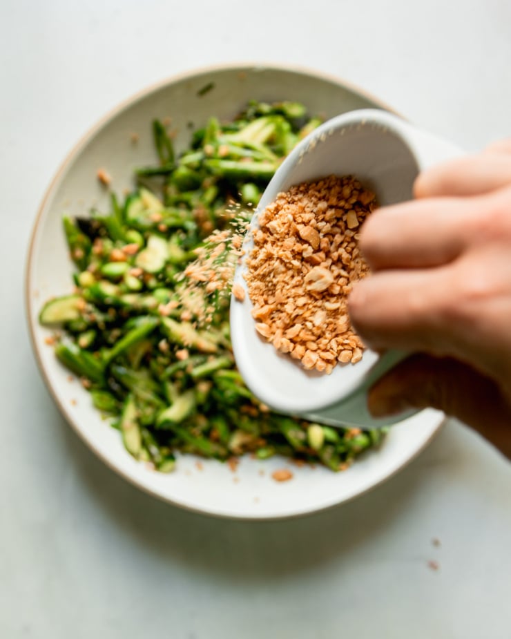 An overhead shot shows a hand emptying a bowl of finely chopped salted peanuts over a salad.
