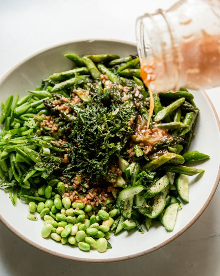 A 3/4 angle shot shows salad dressing being poured over chopped snap peas, cucumber, mint, edamame, and roasted asparagus.