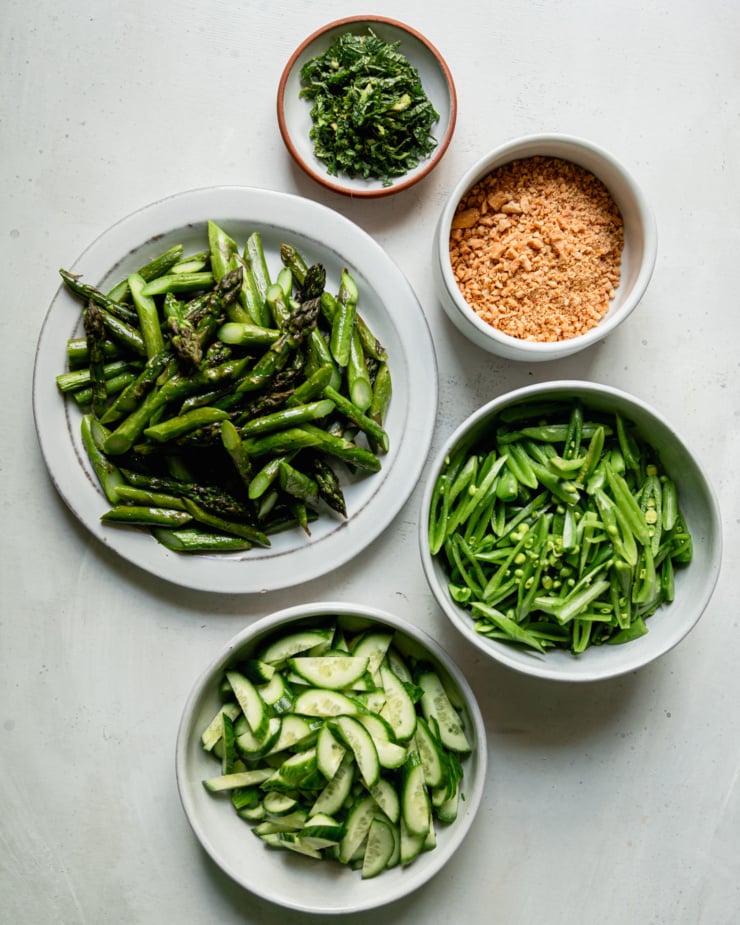An overhead shot shows bowls/plates of the following: sliced mint, finely chopped salted peanuts, sliced snap peas, sliced Persian cucumbers, chopped roasted asparagus.