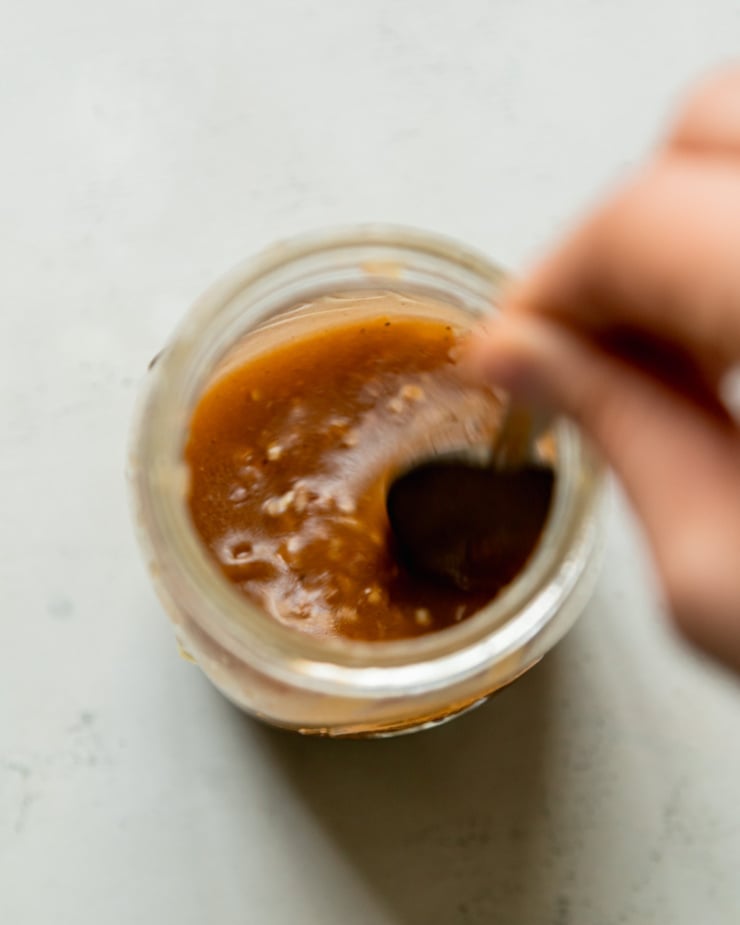 An overhead shot shows a hand using a spoon to stir some ginger lime salad dressing in a jar.