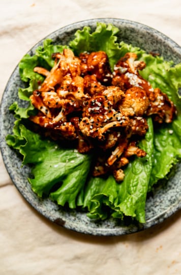 An up close overhead shot shows a singular roasted cauliflower lettuce wrap on an enamelware plate. The cauliflower is topped with glossy sweet chili sauce and sprinkled with sesame seeds.