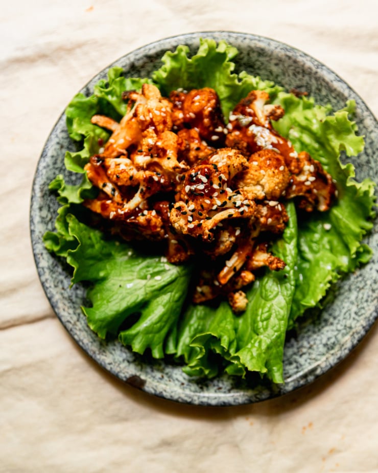 An up close overhead shot shows a singular roasted cauliflower lettuce wrap on an enamelware plate. The cauliflower is topped with glossy sweet chili sauce and sprinkled with sesame seeds.