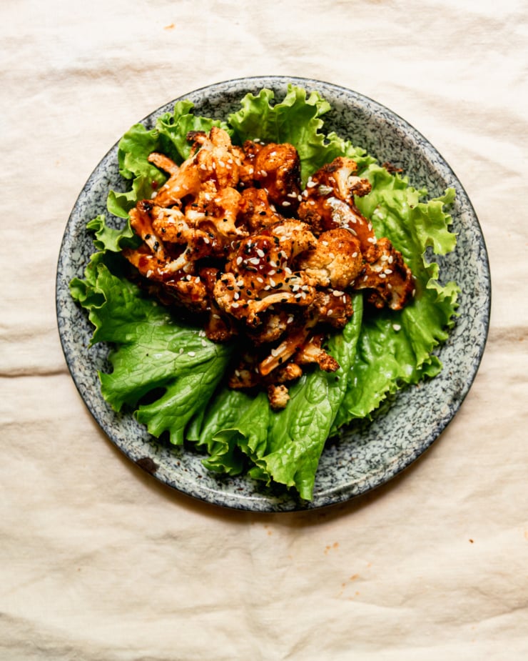 An overhead shot shows a singular roasted cauliflower lettuce wrap on an enamelware plate. The cauliflower is topped with glossy sweet chili sauce and sprinkled with sesame seeds.