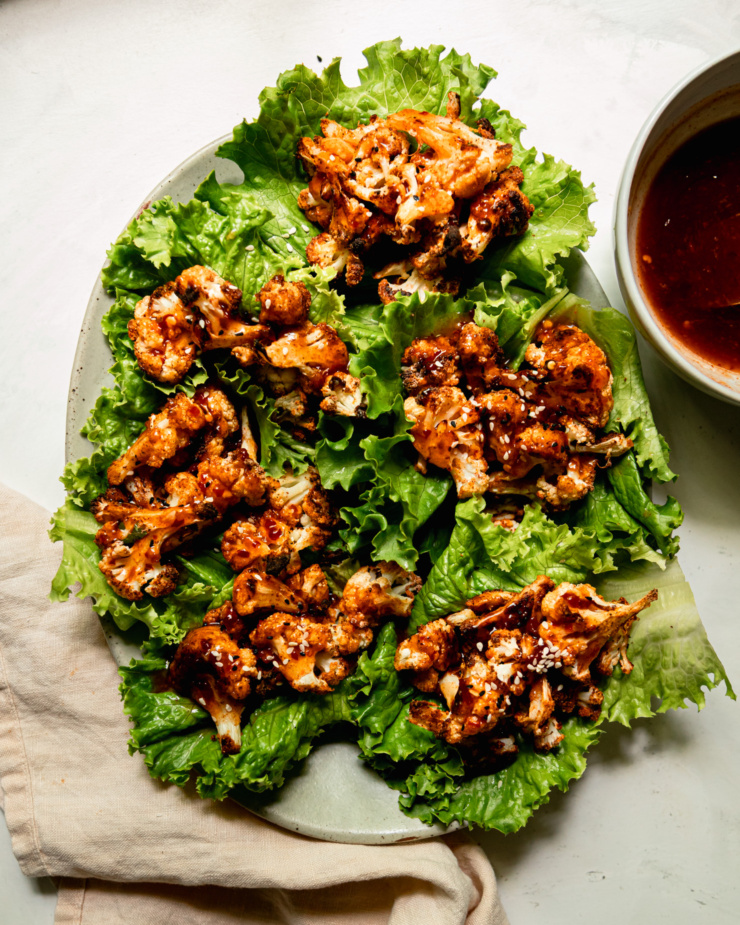 An overhead shot shows a platter of roasted cauliflower lettuce wraps. The cauliflower is drizzled liberally with homemade sweet chili sauce. A bowl of the sauce is seen to the side, slightly out of frame.