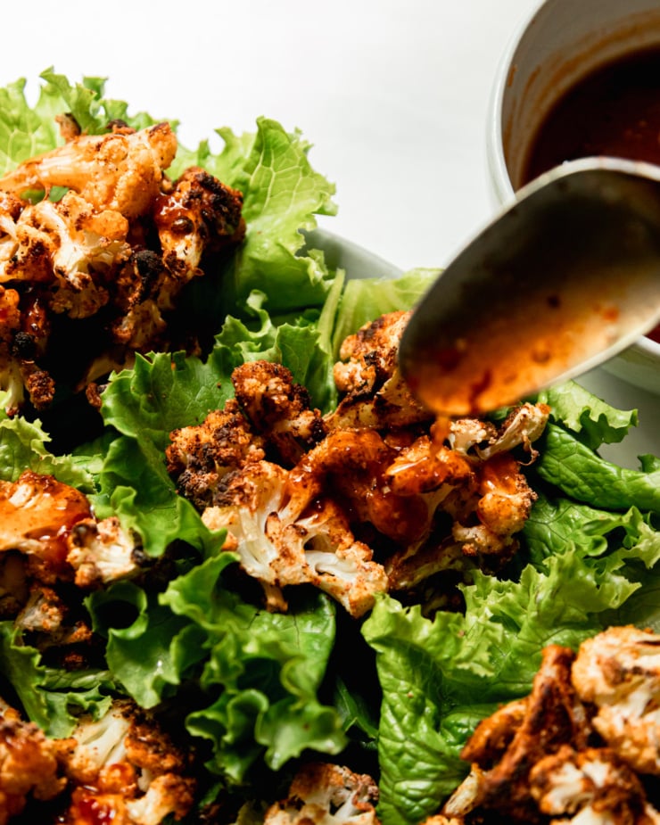An overhead shot shows sweet chili sauce being drizzled onto roasted cauliflower lettuce wraps with a spoon.