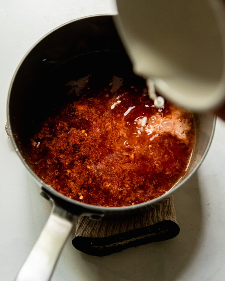 An overhead shot shows a cornstarch slurry being poured into a saucepan of homemade sweet chili sauce.