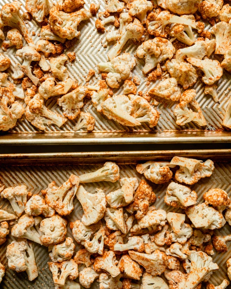 An overhead shot shows cauliflower coated in oil, spices, and cornstarch on 2 baking sheets, prior to being roasted.