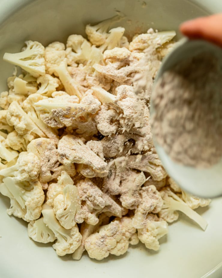 An overhead shot shows a hand sprinkling a mixture of cornstarch and spices over cauliflower florets in a large bowl.