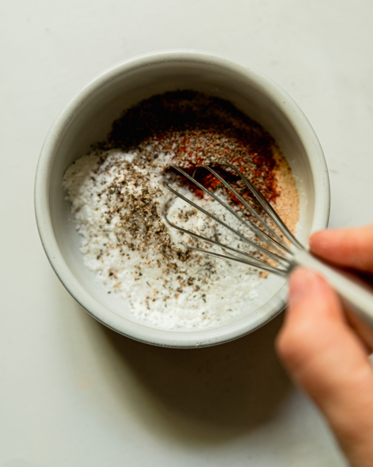 An overhead shot shows a hand using a whisk to bring together, cornstarch, spices, salt and pepper in a small bowl.