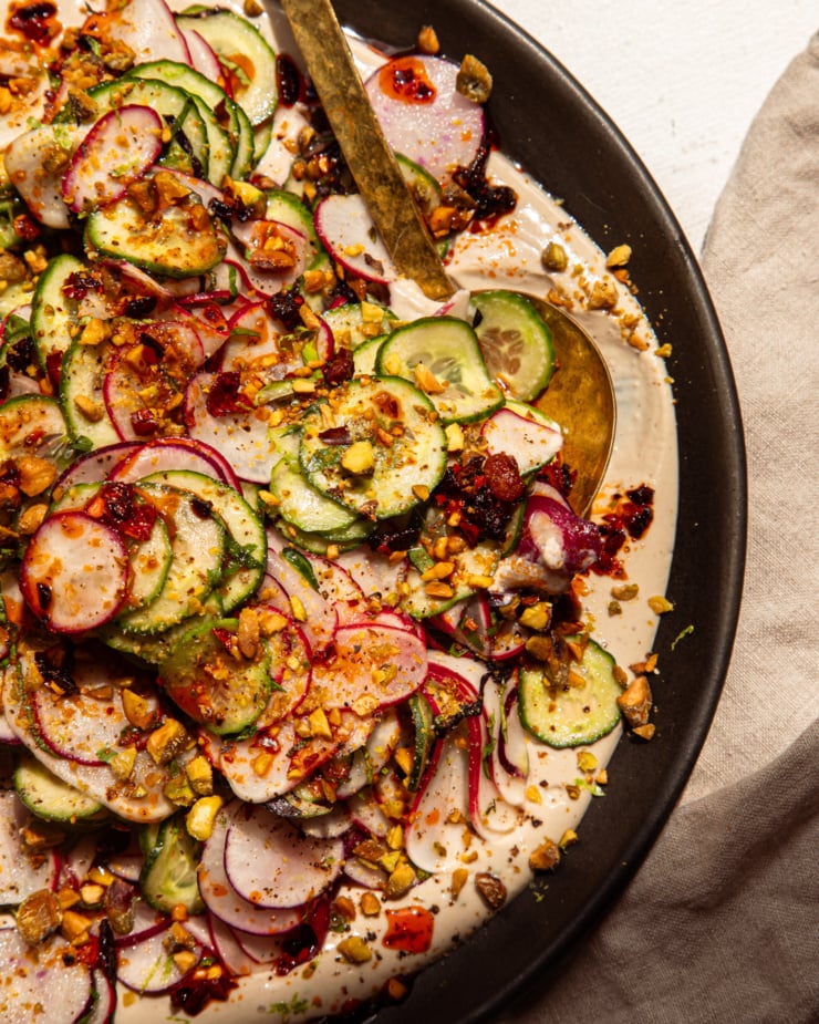 An up close, overhead shot shows a shaved cucumber radish salad on a moat of garlic lime tahini. The salad is garnished with finely chopped pistachios and chili crisp. A serving spoon is tucked into the side of the salad.