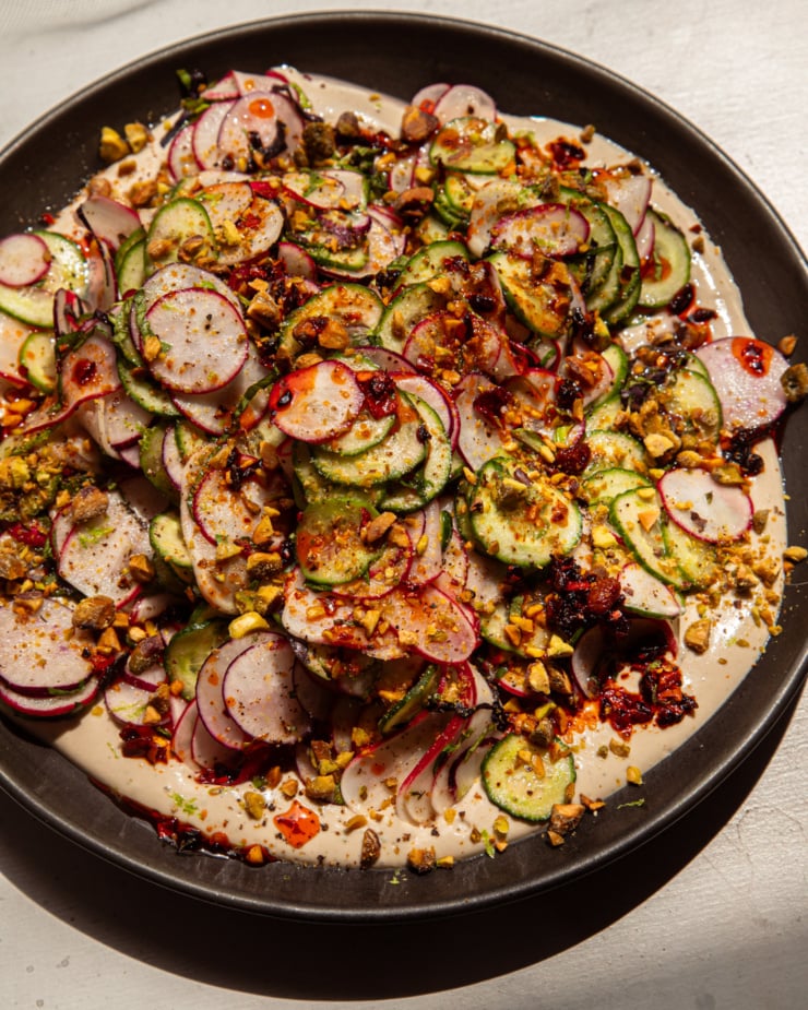 A brightly lit, overhead shot shows a shaved cucumber radish salad on a moat of garlic lime tahini. The salad is garnished with finely chopped pistachios and chili crisp.