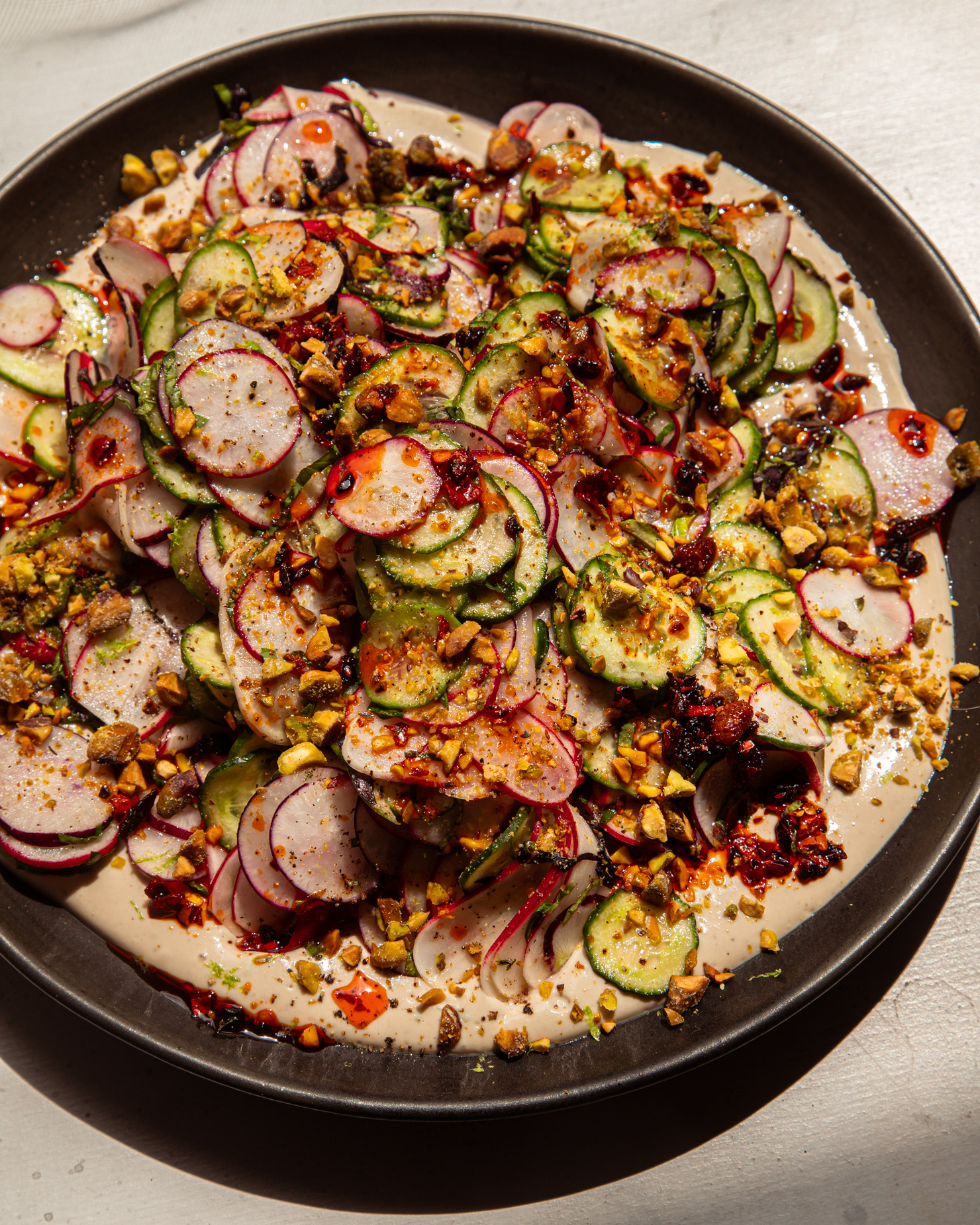 A brightly lit, overhead shot shows a shaved cucumber radish salad on a moat of garlic lime tahini. The salad is garnished with finely chopped pistachios and chili crisp.
