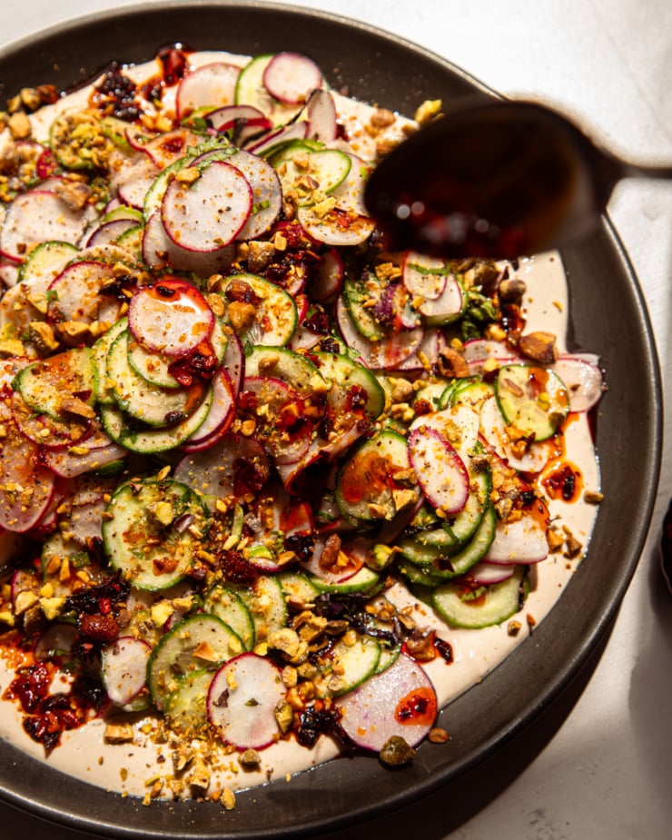 A brightly lit, overhead shot shows a shaved cucumber radish salad on a moat of garlic lime tahini. The salad is garnished with finely chopped pistachios and chili crisp. A spoon is dripping chili crisp onto the salad.