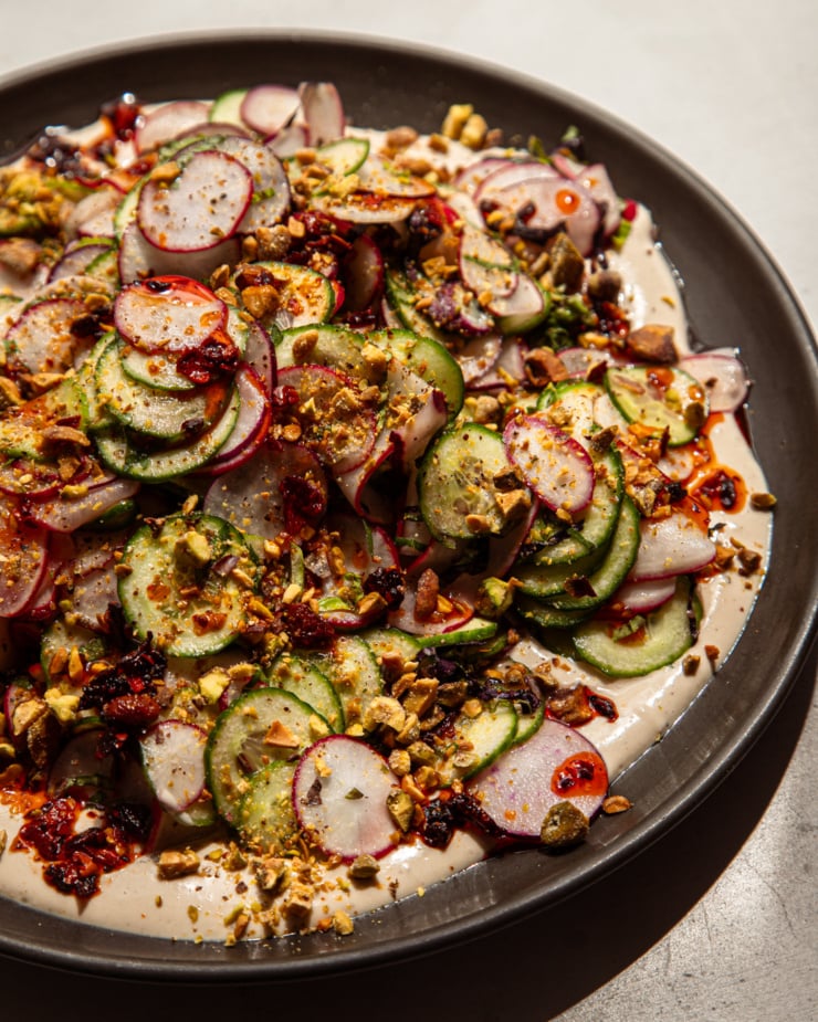 A 3/4 angle shot shows a shaved cucumber radish salad on a moat of garlic lime tahini. The salad is garnished with finely chopped pistachios and chili crisp.