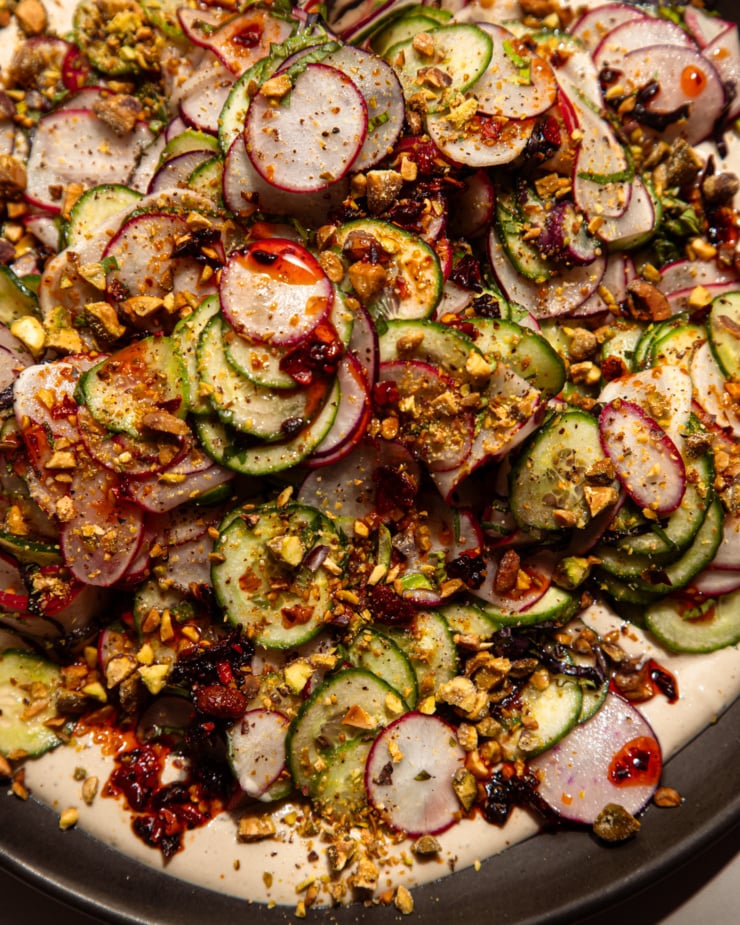 An up close, overhead shot shows a shaved cucumber radish salad on a moat of garlic lime tahini. The salad is garnished with finely chopped pistachios and chili crisp.
