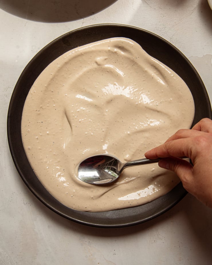 An overhead shot shows a hand using a spoon to spread a tahini-based sauce out on a plate.