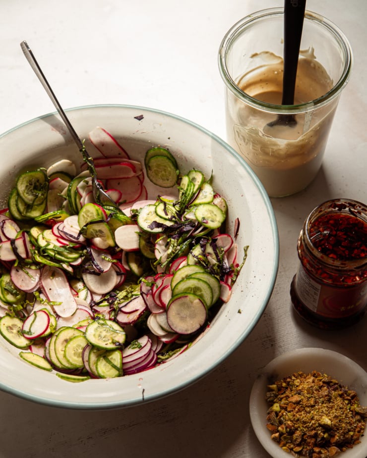 A 3/4 angle shot shows a shaved radish and cucumber salad in a bowl, a tahini-based sauce, a jar of chili crisp, and finely chopped pistachios.