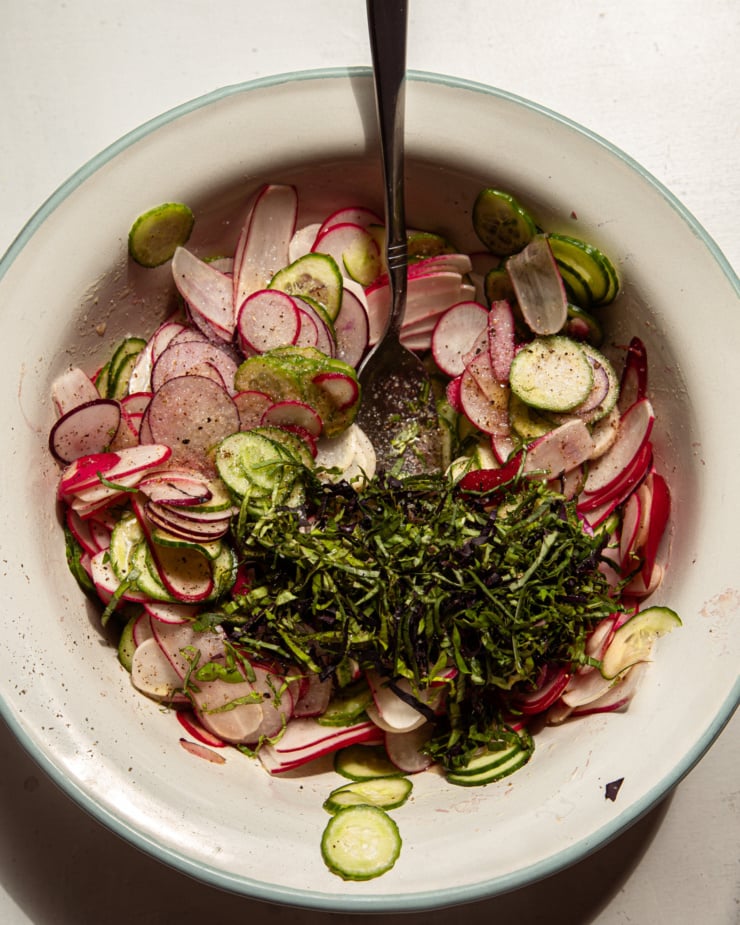 An overhead shot shows a bowl filled with sliced radish, cucumber, and basil.