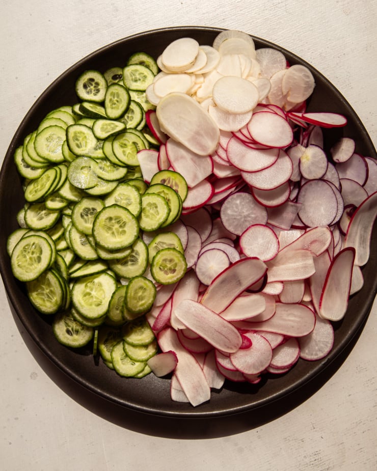 An overhead shot shows a dark plate filled with thinly sliced radishes and cucumbers.
