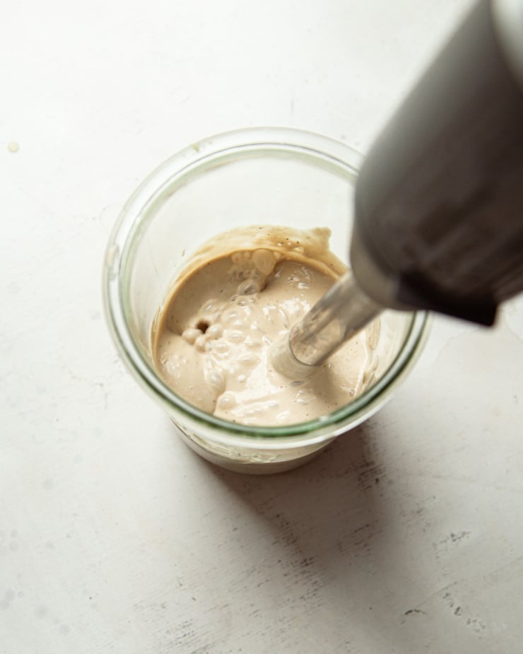 An overhead shot shows a stick blender bringing a tahini dressing together in a jar.