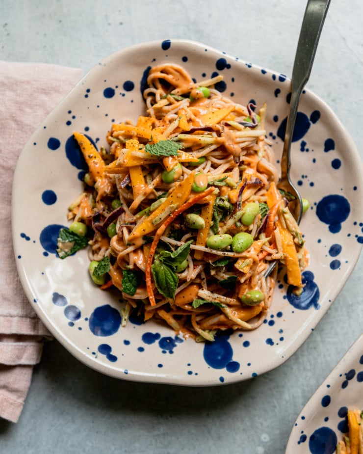 An overhead shot shows an individual serving of mango slaw noodles in a shallot bowl. The noodles feature bagged slaw mix, mango, shelled edamame, and chopped herbs.