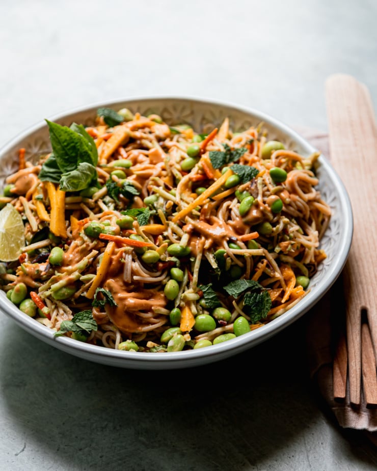 A 3/4 angle shot shows a bowl of mango slaw noodles with shelled edamame and lots of chopped mint and basil. Wooden serving tongs are seen to the side.