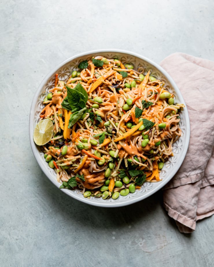 An overhead shot shows a bowl of mango slaw noodles with shelled edamame and lots of chopped mint and basil. A linen napkin is close to the bowl.