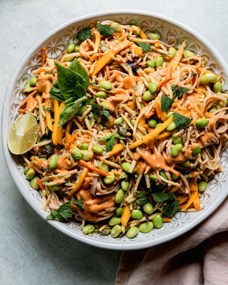 An overhead shot shows a bowl of mango slaw noodles with shelled edamame and lots of chopped mint and basil.