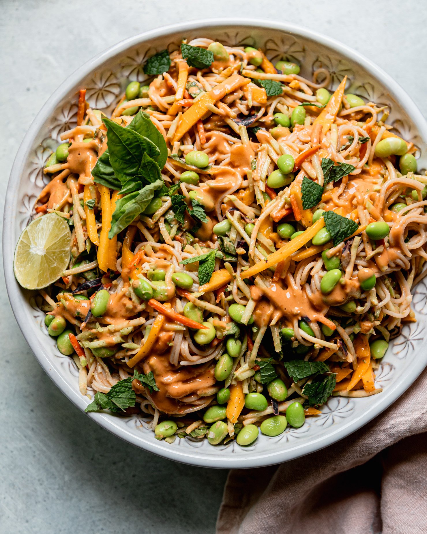 An overhead shot shows a bowl of mango slaw noodles with shelled edamame and lots of chopped mint and basil.