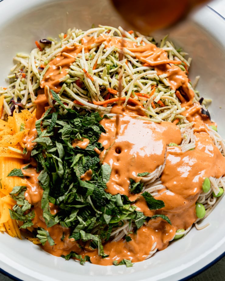 An overhead shot shows peanut sauce being poured over noodles, slaw mix, mango, and chopped herbs.