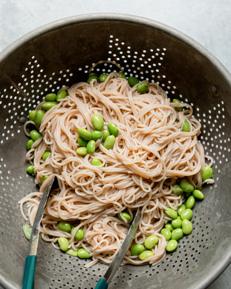 An overhead show shows a colander filled with cooked gluten-free cappellini and shelled edamame.