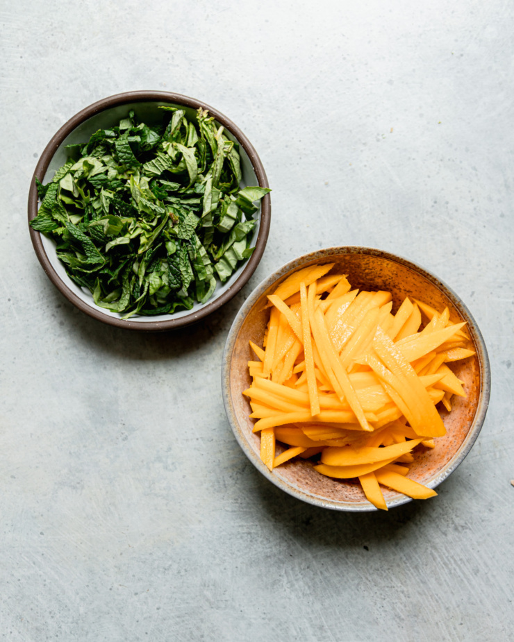 An overhead shot shows a bowl of chopped herbs and a bowl of julienned mango.