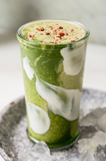 A head-on shot shows a matcha banana smoothie in a glass. The top is garnished with crushed toasted pecans. Through the glass you can see swirls of light and deep green and also white.