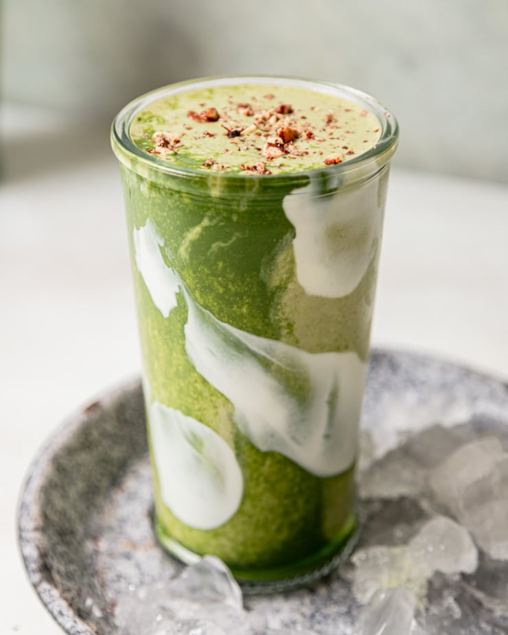 A head-on shot shows a matcha banana smoothie in a glass. The top is garnished with crushed toasted pecans. Through the glass you can see swirls of light and deep green and also white.