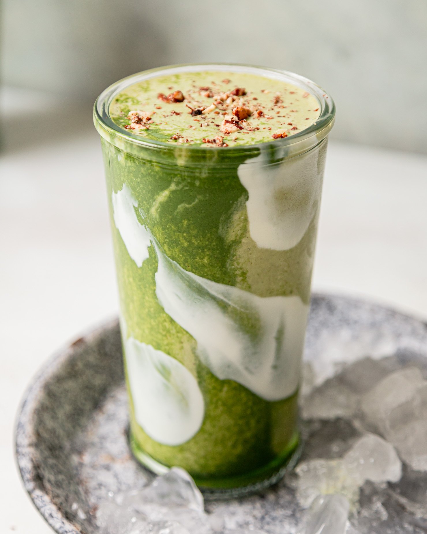 A head-on shot shows a matcha banana smoothie in a glass. The top is garnished with crushed toasted pecans. Through the glass you can see swirls of light and deep green and also white.