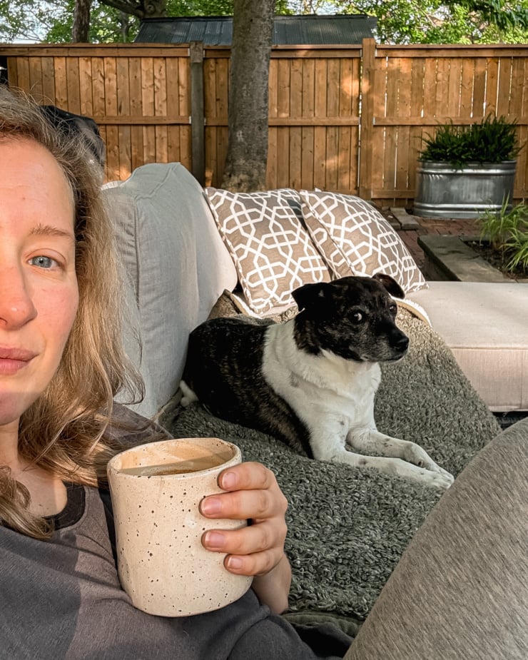 A selfie shows a woman enjoying a coffee outside along with her dog in the background.