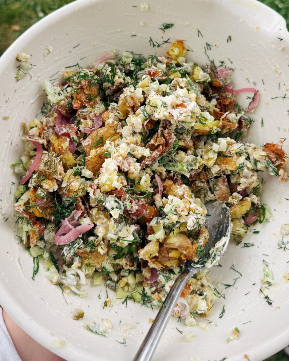 An overhead shot shows a crispy smashed potato salad in a bowl with pickles, pickled red onion, celery, and creamy dill dressing.