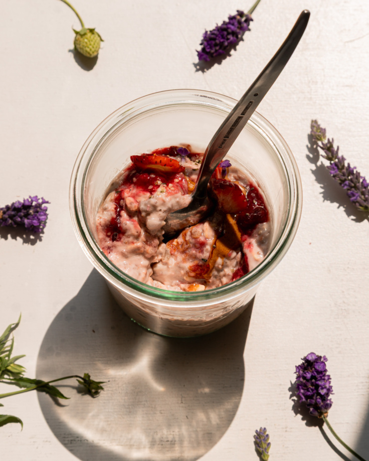An overhead shot shows a serving of strawberry rhubarb overnight oats in a glass jar. The top is garnished with fresh strawberries, almond butter, compote, hemp seeds, and lavender flowers. The jar is framed by lavender flowers on the stem.