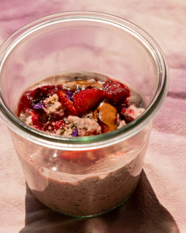 An up close 3/4 angle shot shows a serving of strawberry rhubarb overnight oats in a glass jar. The top is garnished with fresh strawberries, almond butter, compote, hemp seeds, and lavender flowers.