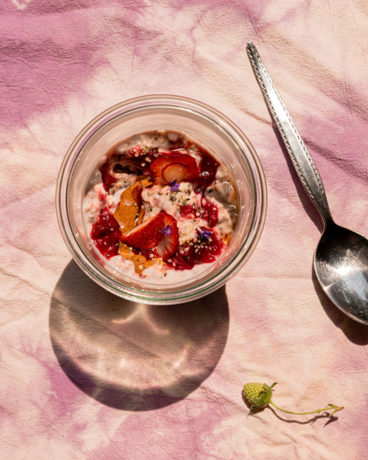 An overhead shot shows a serving of strawberry rhubarb overnight oats in a glass jar. The top is garnished with fresh strawberries, almond butter, compote, hemp seeds, and lavender flowers. A spoon is nearby, gleaming in the bright sun light. A teeny green strawberry on the stem is seen in the bottom corner.