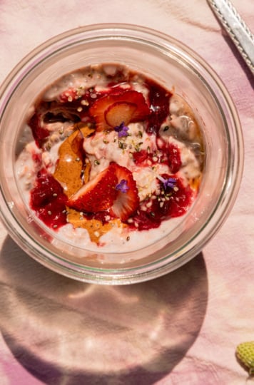 An up close, overhead shot shows a serving of strawberry rhubarb overnight oats in a glass jar. The top is garnished with fresh strawberries, almond butter, compote, hemp seeds, and lavender flowers. A teeny green strawberry on the stem is seen in the bottom corner.