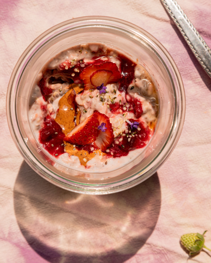 An up close, overhead shot shows a serving of strawberry rhubarb overnight oats in a glass jar. The top is garnished with fresh strawberries, almond butter, compote, hemp seeds, and lavender flowers. A teeny green strawberry on the stem is seen in the bottom corner.