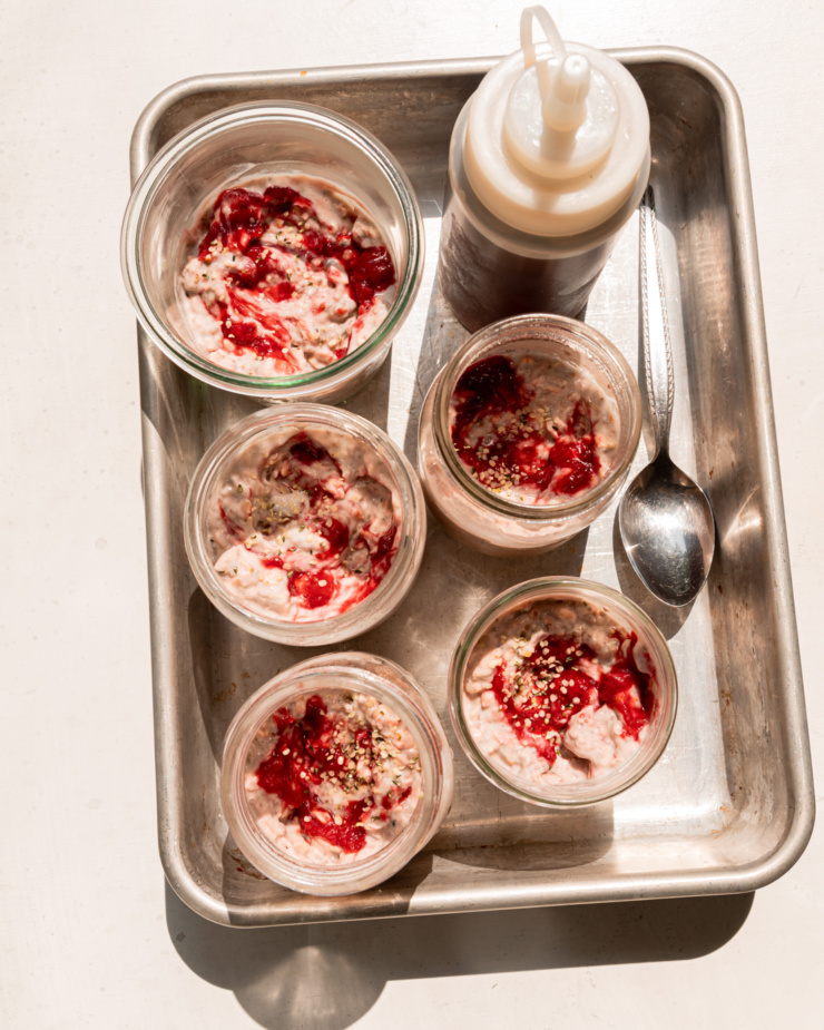 An overhead shot shows a steel tray with 5 jars of rhubarb strawberry overnight oats. A squeeze bottle of maple syrup is also in the tray. The photo is taken in direct sunlight.
